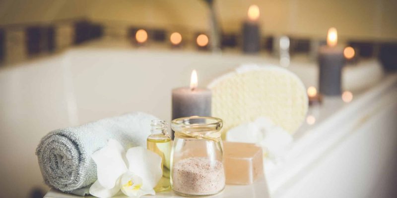 White ceramic tray with home spa supplies in home bathroom for relaxing rituals. Candlelight, salt soap bar, bath salt in jar, massage, bath oil in bottle, blue rolled towel, natural sponge.